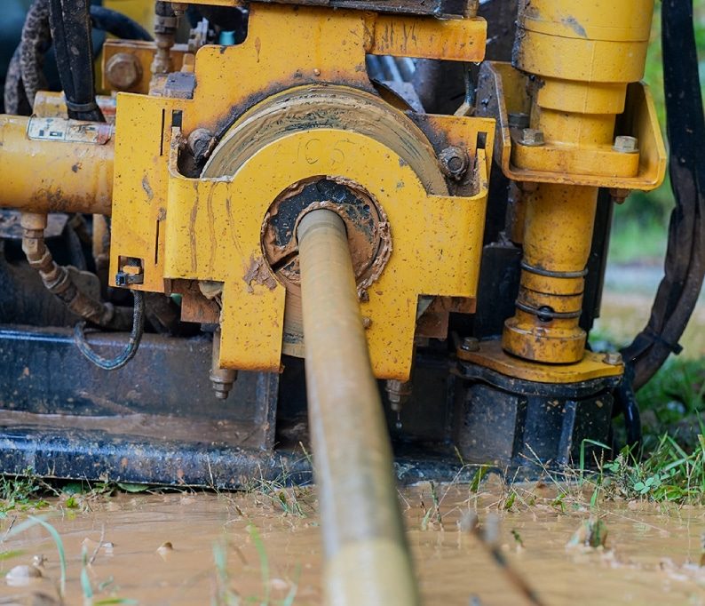 A close-up view of a horizontal directional drilling machine operating in muddy conditions, with a metal drill rod extending from the mechanism.