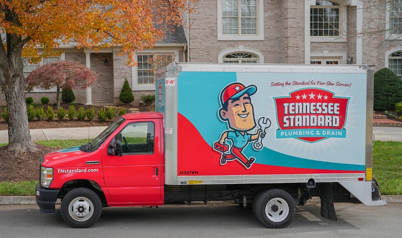 A red and white Tennessee Standard moving truck is parked on a residential street in front of brick homes with fall foliage in the background.