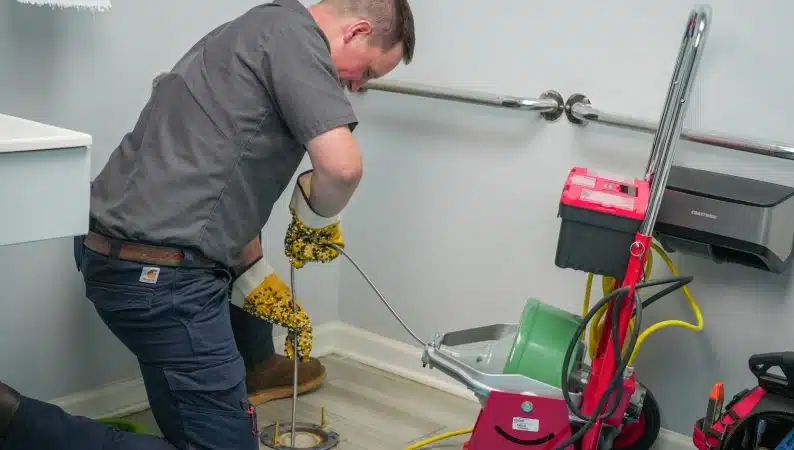 A plumber kneels on the floor using a drain snake to unclog a drain, with plumbing equipment and tools around him in a utility room.