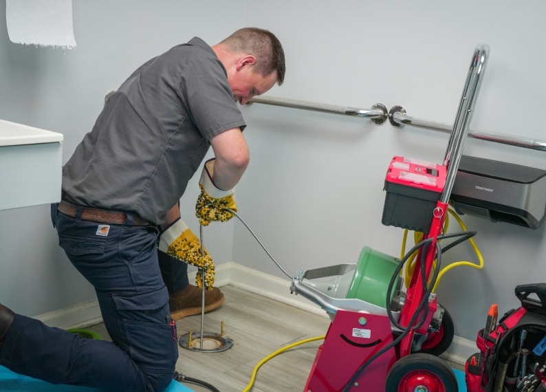A plumber kneels on the floor using a drain snake to unclog a drain, with plumbing equipment and tools around him in a utility room.