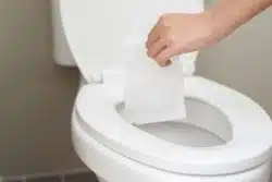 A hand hovers over a toilet, about to drop a piece of white toilet paper into the open bowl. The scene suggests a bathroom setting.