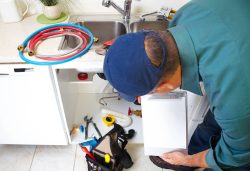 A plumber in a blue shirt and cap examines under a kitchen sink with tools and hoses nearby. He holds a clipboard while evaluating the repair.