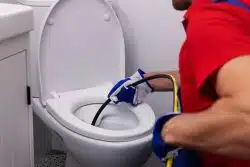 A plumber in a red shirt and blue gloves uses a drain snake to unclog a white toilet in a bright bathroom, conveying a sense of focus and practicality.