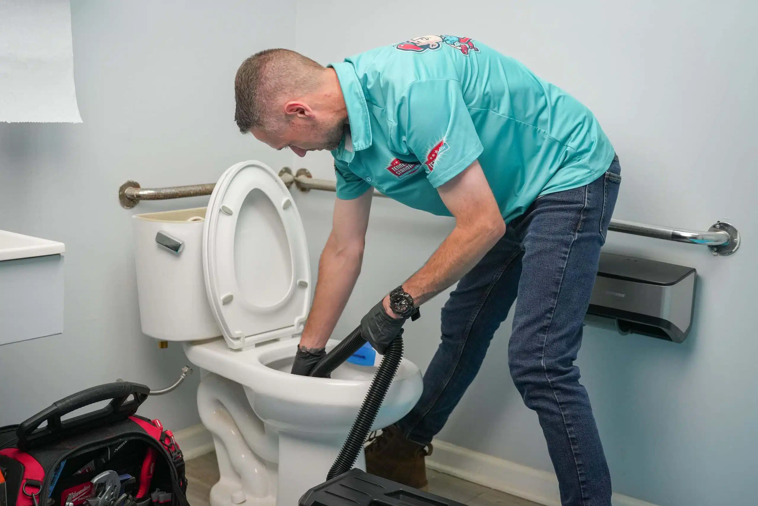 A plumber in a teal shirt and jeans unclogs a white toilet using a tool. A tool bag is on the floor in a small bathroom with a paper towel dispenser.