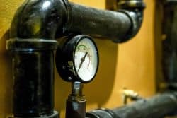 Close-up of a pressure gauge on black industrial pipes against a yellow wall. The gauge needle is in the green zone, indicating normal pressure.
