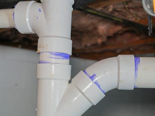 Close-up of white PVC plumbing pipes with purple primer at the joints, installed in a basement with exposed insulation and wooden beams.