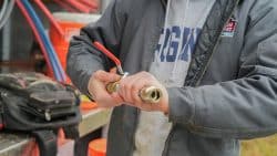 A person in a gray jacket attaches a nozzle to a hose, standing next to a black tool bag and orange equipment—possibly troubleshooting sudden drops in water pressure.