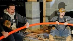 Two workers sit on a dirt floor, pulling a flexible orange pipe between them in a basement area with concrete blocks and wooden beams—addressing plumbing issues after renovations.
