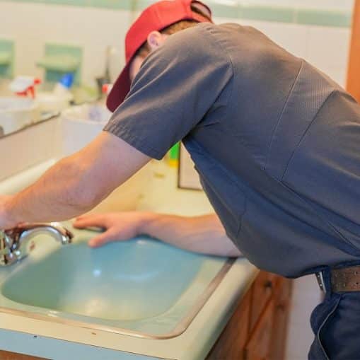 A person wearing a red cap and work uniform is fixing or inspecting a bathroom sink faucet, possibly addressing sudden drops in water pressure.