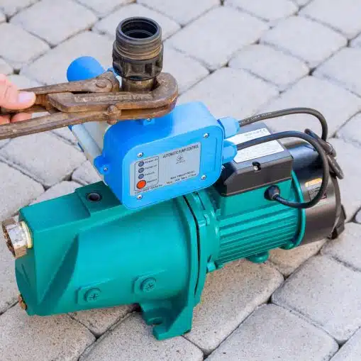 A teal automatic water pump with a blue pressure controller mounted on top, sitting on a paver stone surface while a hand tightens a fitting with a wrench.