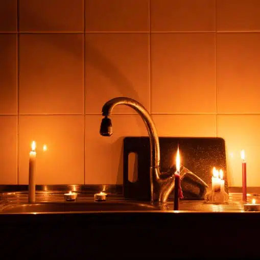 A kitchen sink lit by two candles with a paper displaying a sad face propped against the faucet; the dimly lit setting hints at plumbing risks during power outages.