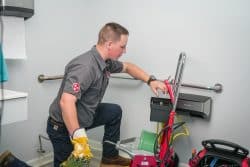 A plumber kneels on the floor using a commercial drain cleaning machine in a bathroom equipped with handrails and cleaning supplies.