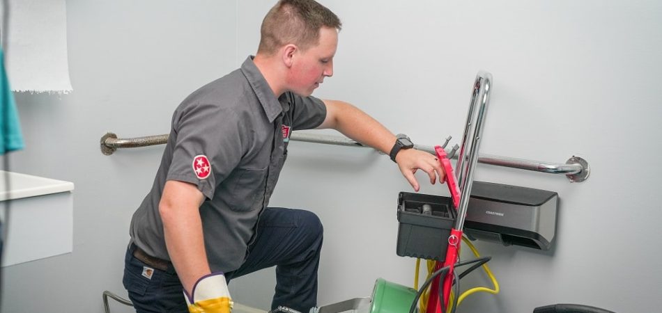 A plumber kneels on the floor using a commercial drain cleaning machine in a bathroom equipped with handrails and cleaning supplies.