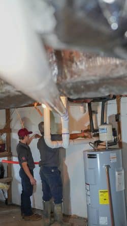 Two workers in uniforms install white PVC pipes in a basement near a hot water heater and furnace, with tools and equipment visible—demonstrating expert commercial drain cleaning services.