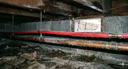 Red and copper pipes run along the ceiling of a crawl space with exposed wooden beams, cinder block walls, and a small window letting in some light.