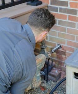 A person with short hair is kneeling and working on outdoor plumbing near a brick wall, using tools and equipment for Backflow Testing in Knoxville.
