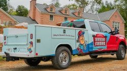 A Tennessee Standard Plumbing & Drain service truck is parked on a gravel driveway in front of a large brick house under construction, ready to tackle outdoor drain unclogging and other plumbing needs.