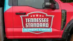 Red pickup truck door with "Tennessee Standard Plumbing & Drain" logo and white stars, viewed from the side on a rainy day.