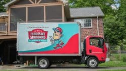 A red and teal Tennessee Standard Plumbing & Drain truck is parked in a driveway beside a brick house with a raised wooden porch, ready to service drainage systems in Knoxville.