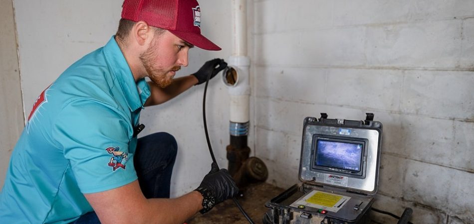 A technician in a teal shirt and red cap inspects a pipe for drain cleaning in Knoxville, using a camera cable and viewing the results on a monitor in a utility room with concrete walls.