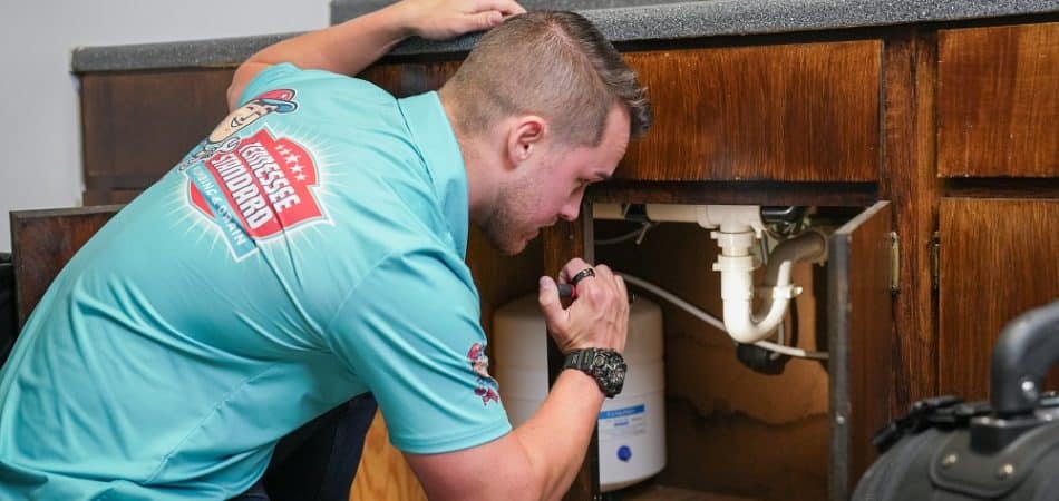 A person wearing a blue shirt uses a flashlight to inspect plumbing under a kitchen sink cabinet, checking for plumbing issues after renovations.