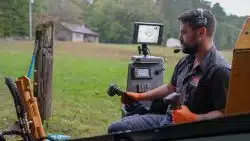 A man wearing orange gloves and a headset operates machinery with control sticks, sitting in a cab with display screens, managing tasks affected by seasonal rainfall in Knoxville that impacts drain performance in the outdoor rural setting.