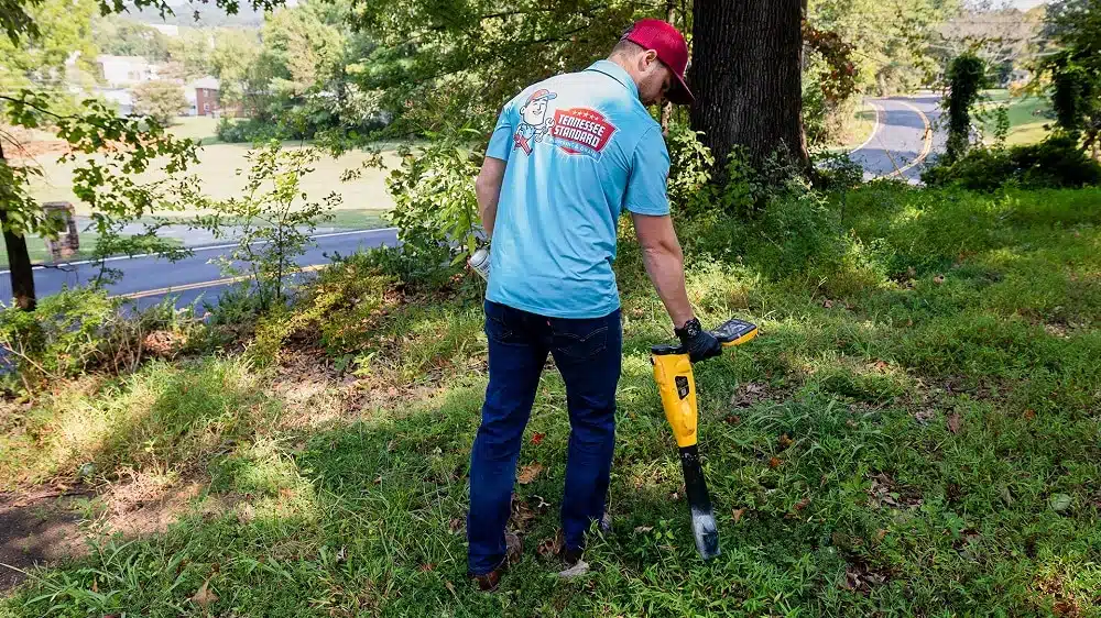 A person in a blue shirt and red cap uses a yellow metal detector on a grassy hillside near a road and trees, perhaps searching for hidden pipes or signs of plumbing problems.