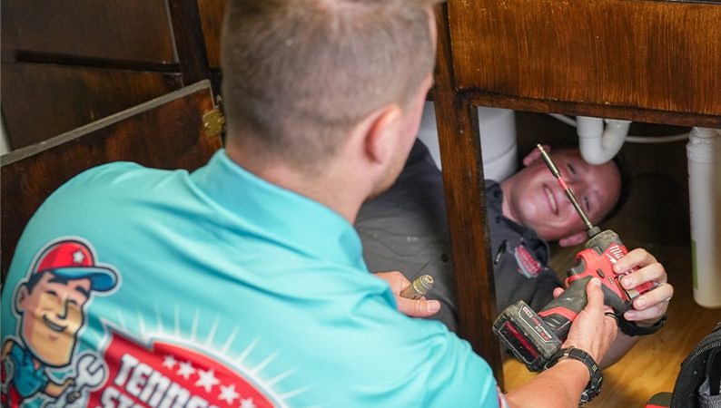 Two plumbers work under a kitchen sink; one holds a power drill while the other lies inside the cabinet. The visible shirt reads
