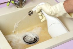 A person in rubber gloves sprays an alkaline cleaning solution onto a dirty kitchen sink.