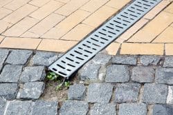 A metal drainage grate runs diagonally between yellow tiles and gray cobblestones, with green plants growing through a crack in the cobblestone—a reminder of the importance of regular outdoor drain unclogging.