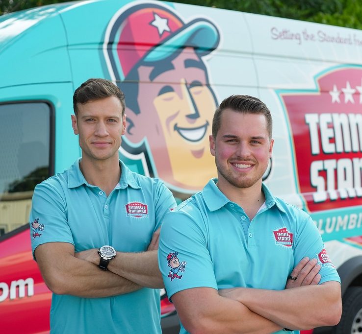 Bobby and Kelton Balka in light blue shirts stand smiling with arms crossed in front of a van displaying the Tennessee Standard logo and colorful graphics.