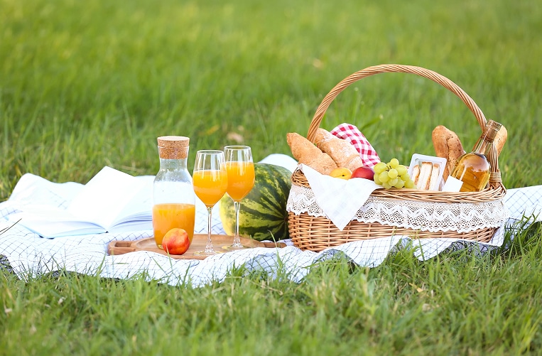 A picnic setup on lush grass features a wicker basket with bread, grapes, and wine