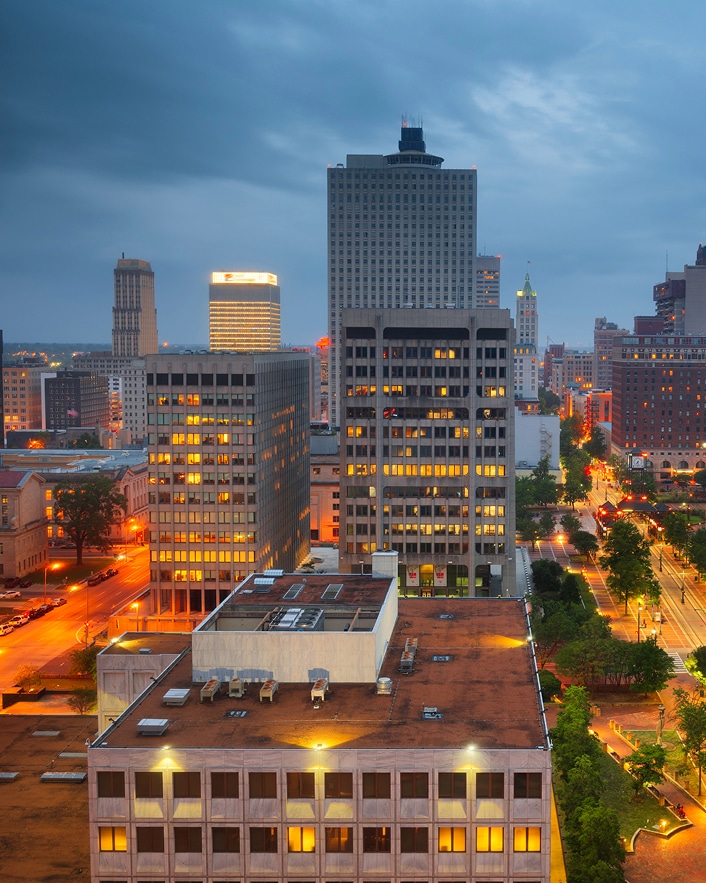 Cityscape at dusk with glowing lights in office buildings and street lamps, under a cloudy sky in Alcoa