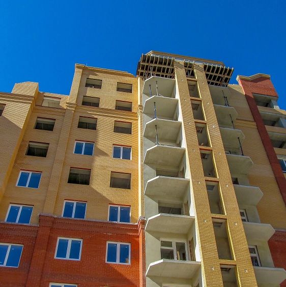 A multi-story apartment building with red and yellow exterior walls, balconies, and windows, viewed from below against a clear blue sky.
