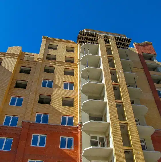 A multi-story apartment building with red and yellow exterior walls, balconies, and windows, viewed from below against a clear blue sky.