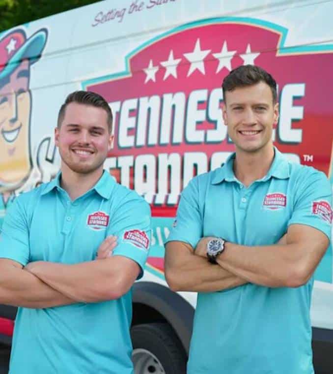 Bobby and Kelton Balka wearing a light blue shirts stand smiling with arms crossed in front of a van displaying the Tennessee Standard logo and colorful graphics.