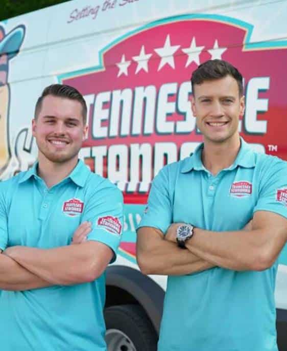Two smiling men in blue uniform shirts stand with arms crossed in front of a vehicle with