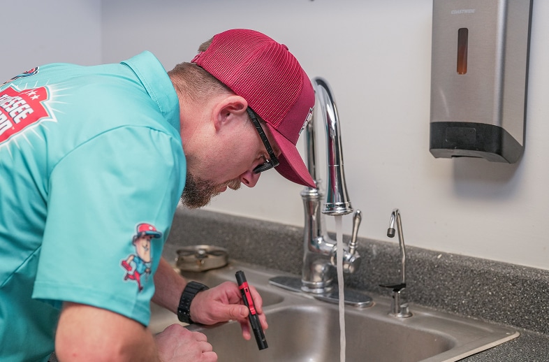 A person wearing a teal shirt and red cap inspects a sink with running water using a flashlight in a kitchen or utility room.