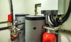 A mechanical room with two large water tanks, a wall-mounted boiler, various pipes, valves, gauges, and a red expansion tank—ideal for those seeking a reliable water heater in Oak Ridge.