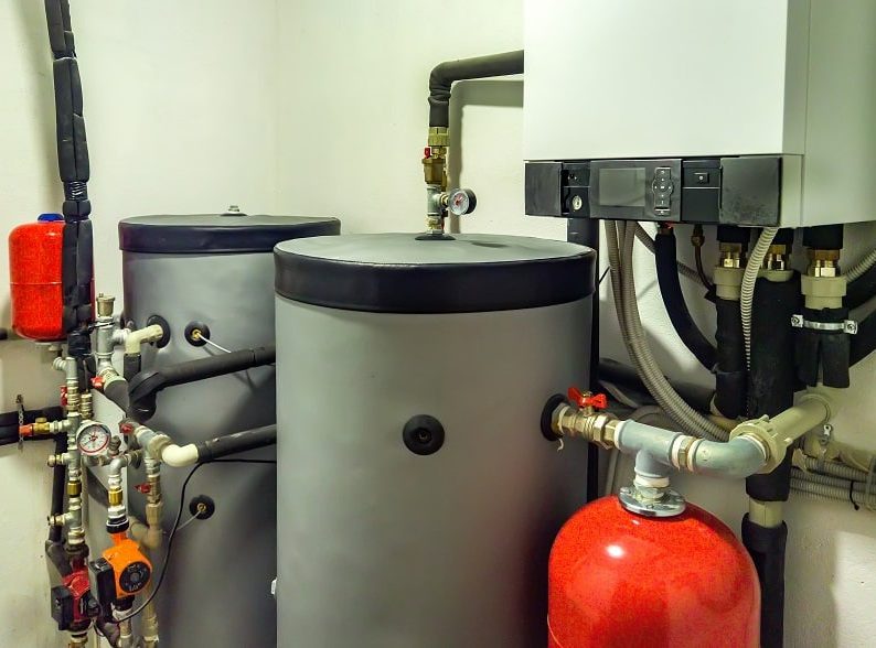 A mechanical room with two large water tanks, a wall-mounted boiler, various pipes, valves, gauges, and a red expansion tank—ideal for those seeking a reliable water heater in Oak Ridge.
