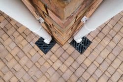 Two white downspouts attached to a brick corner wall, draining into black grates set in tan stone paving—ideal for easy outdoor drain unclogging.