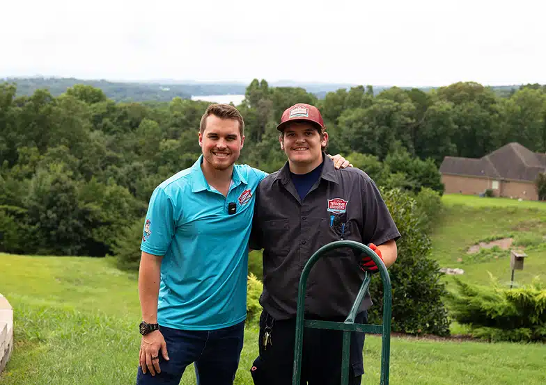 Kelton and his mate pose and smile outdoors on a grassy lawn, with trees and houses in the background. One wears a light blue shirt, the other a dark uniform and cap, standing next to a green dolly.