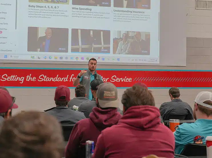 Kelton Balka gives a presentation to a seated audience in a classroom, with financial topics displayed on a large screen behind him.