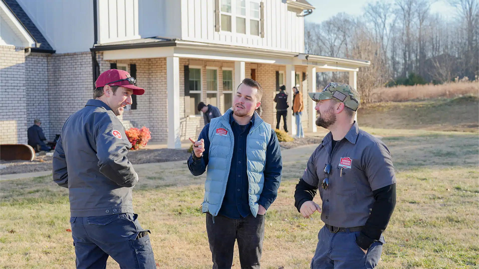 Three men stand outside a house talking; two wear matching work uniforms while the third wears a blue vest.