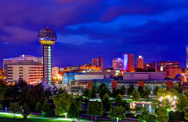 Cityscape at dusk with a tall observation tower featuring a spherical top, surrounded by modern buildings and trees illuminated by city lights.