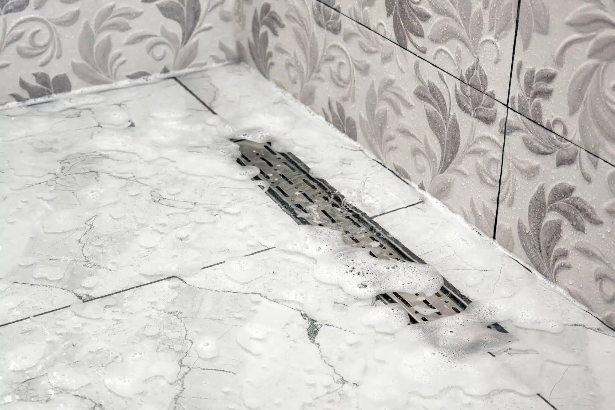 Soapy water on a tiled bathroom floor drains through a rectangular metal grate near the corner, with decorative leaf-patterned wall tiles visible.