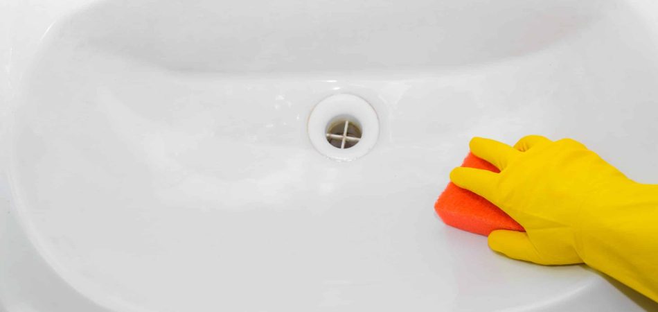 A person wearing a yellow rubber glove cleans a white sink with an orange sponge, showcasing the importance of regular drain cleaning in Knoxville.