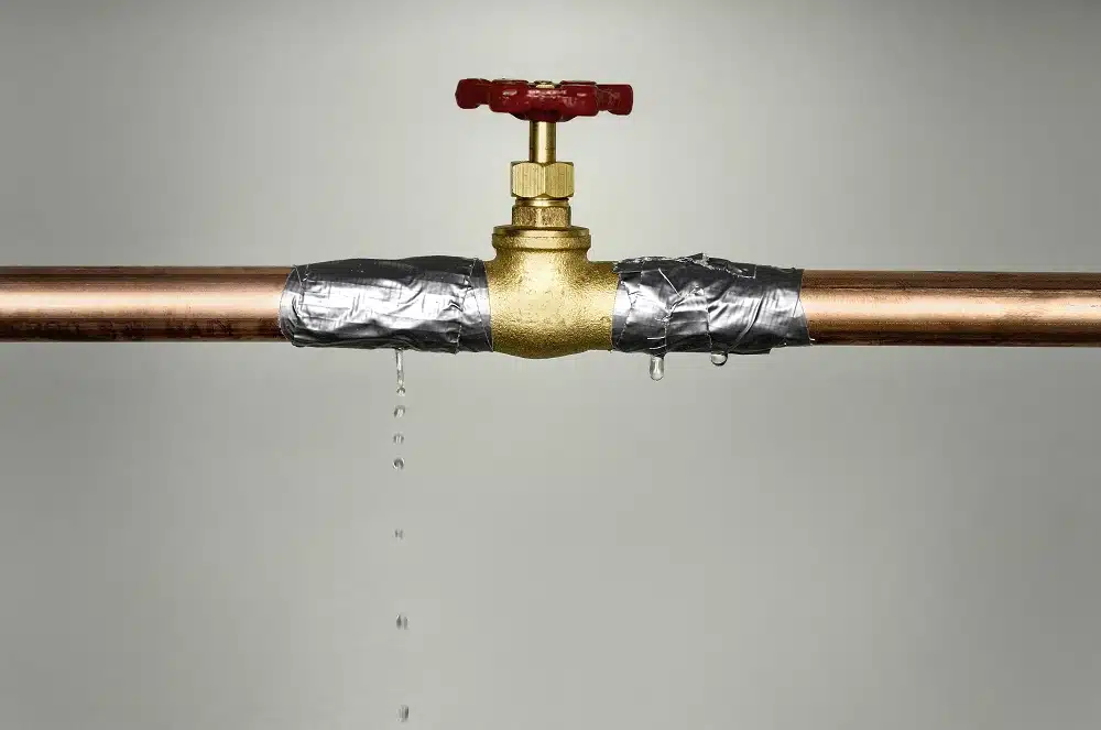 A brass valve on a copper pipe wrapped with duct tape struggles against high water pressure, leaking droplets onto a plain background.