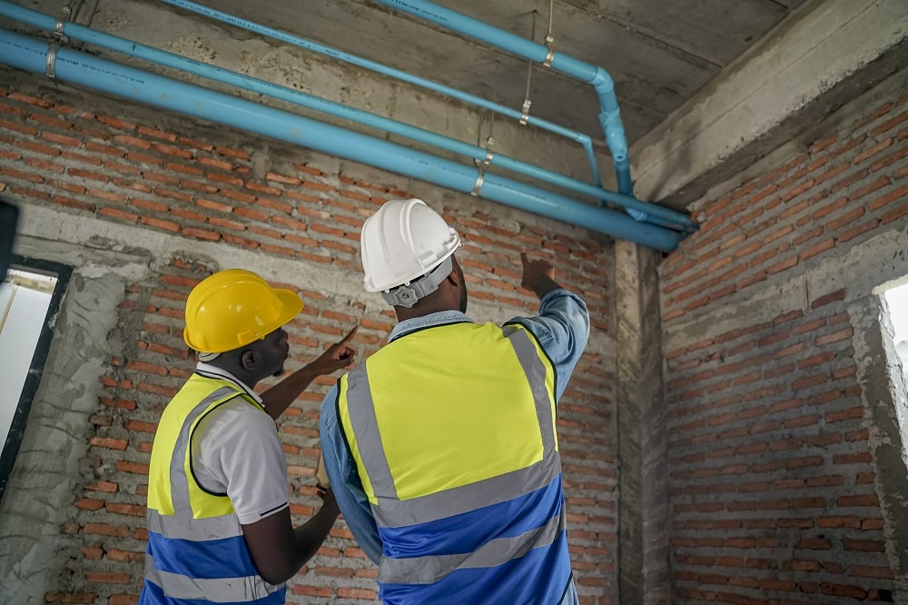 Two construction workers, wearing safety helmets and vests, examine blue plumbing pipes installed in a building. They appear focused on ensuring proper installation and compliance with standards.