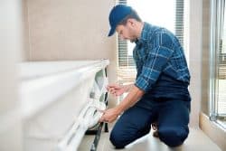 Man in work clothes and cap kneeling on bathroom floor, inspecting or fixing the bathtub drain beneath a tub.